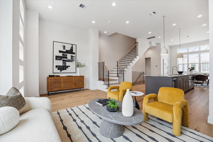 This modern living space features an open floor plan with high ceilings and ample natural light. It includes a cozy seating area with a stylish sofa and two mustard-colored chairs, a sleek kitchen with an island, and contemporary decor. The wooden flooring and staircase add warmth, while large windows enhance the spacious feel.