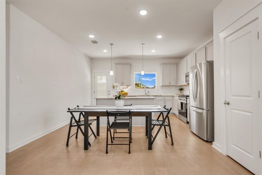 This photo shows a modern kitchen and dining area with light wood floors, white cabinets, and stainless steel appliances. A central island with pendant lighting complements the sleek design, and a large window adds natural light.