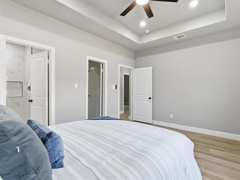 Bedroom with light wood finished floors, a tray ceiling, connected bathroom, a ceiling fan, and recessed lighting Bedroom with light wood finished floors, a tray ceiling, connected bathroom, a ceiling fan, and recessed lighting