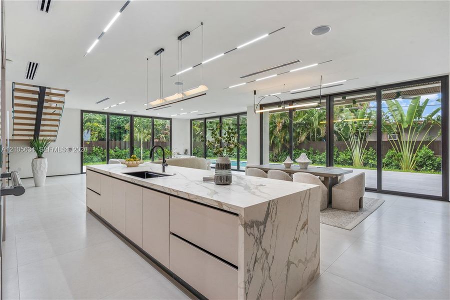 Kitchen featuring a large central island with waterfall-edge stone countertops, custom flat-panel cabinetry, matte black hardware, floor-to-ceiling multi-panel sliding glass doors, and recessed linear LED ceiling lighting