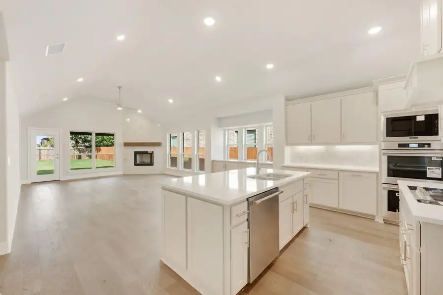 Open kitchen with white cabinets, center island, stainless appliances, and vaulted ceiling open to living area with fireplace.