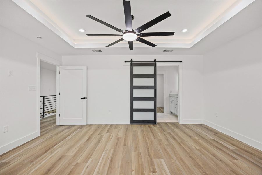 Different angle of primary bedroom featuring a barn door, light wood-style flooring, ensuite bath, a raised ceiling, and recessed lighting