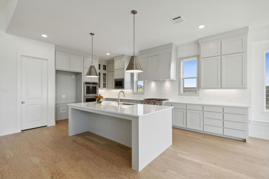 Modern kitchen with a large central island, light wood-finish flooring, white shaker-style cabinetry, and stainless steel pendant lighting Modern kitchen with a large central island, light wood-finish flooring, white shaker-style cabinetry, and stainless steel pendant lighting
