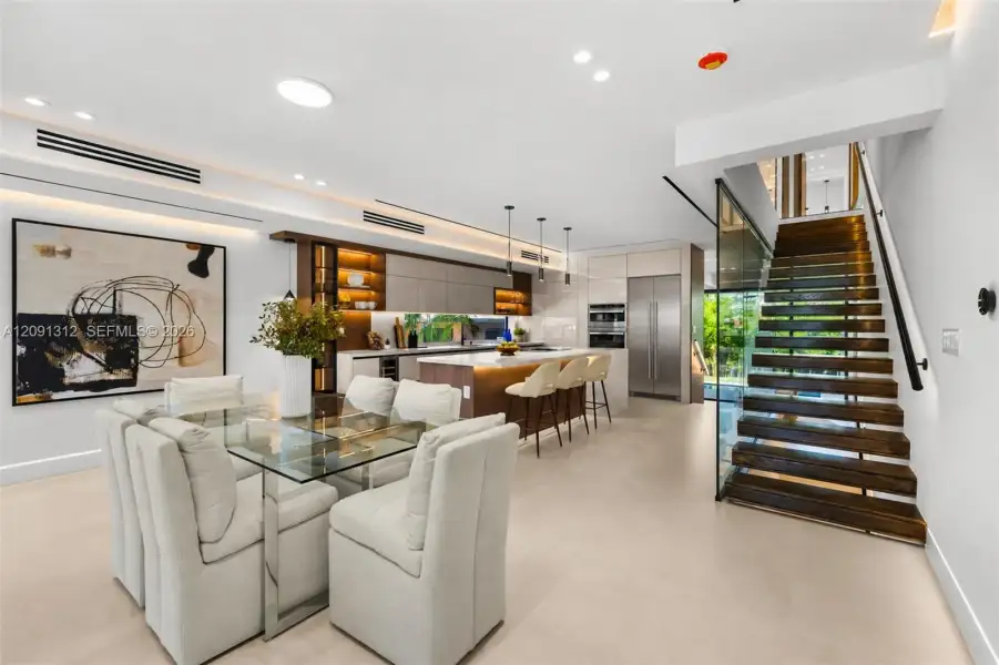 Modern kitchen featuring a stone-topped island with integrated cabinetry, built-in stainless steel appliances, recessed linear lighting, and a floating wood staircase with glass paneling Modern kitchen featuring a stone-topped island with integrated cabinetry, built-in stainless steel appliances, recessed linear lighting, and a floating wood staircase with glass paneling