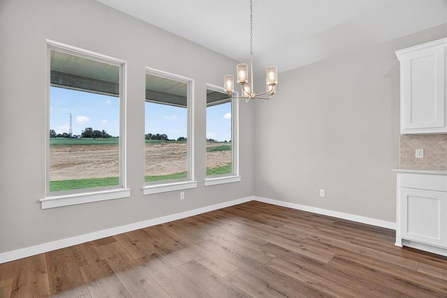 Interior space featuring three rectangular windows, wood-finish flooring, a contemporary chandelier, white baseboards, and built-in white cabinetry with a patterned backsplash Interior space featuring three rectangular windows, wood-finish flooring, a contemporary chandelier, white baseboards, and built-in white cabinetry with a patterned backsplash