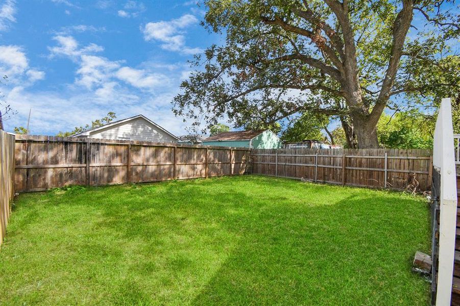 This is a spacious and well-maintained backyard with a wooden privacy fence and a lush green lawn, featuring a clear sky above and part of a house with a balcony visible to the left.