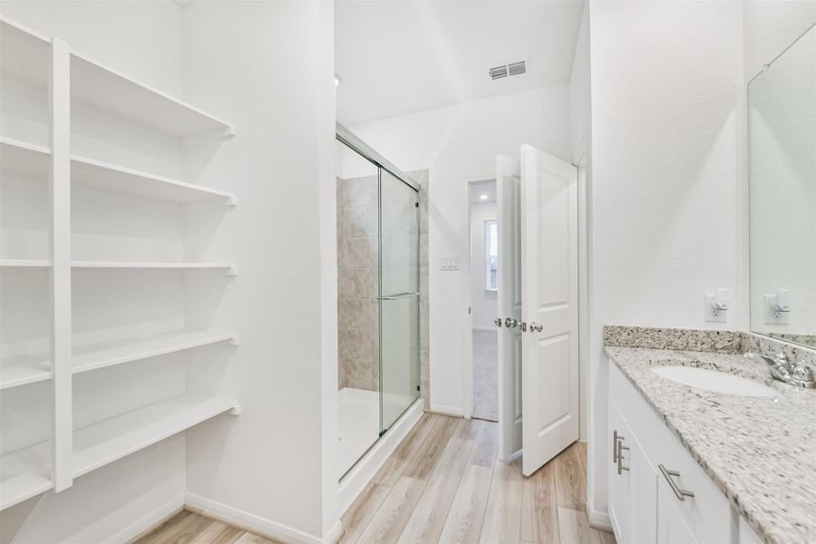 Another view of the primary bathroom showcases a bright, modern bathroom with a glass-enclosed shower, granite countertops, ample shelving, and light wood flooring, leading into the primary bedroom.