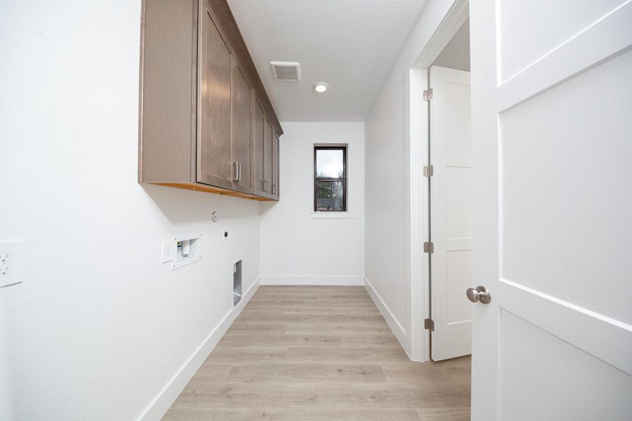 This bright and functional laundry room is conveniently located right off the primary walk-in closet and is ready for your appliances. It features clean white walls, trim, cabinets, window, and laminate flooring.