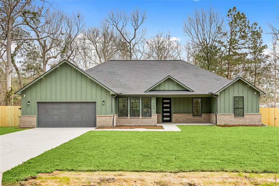 Modern single-story home with a dark green exterior and brick accents, featuring a spacious front lawn. It includes a two-car garage and a stylish front entrance with large windows. The property is bordered by a wooden privacy fence, surrounded by mature trees.