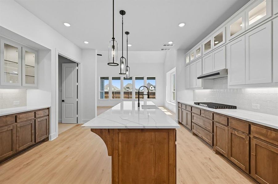 Kitchen featuring tasteful backsplash, glass insert cabinets, brown cabinets, light stone counters, and recessed lighting Kitchen featuring tasteful backsplash, glass insert cabinets, brown cabinets, light stone counters, and recessed lighting