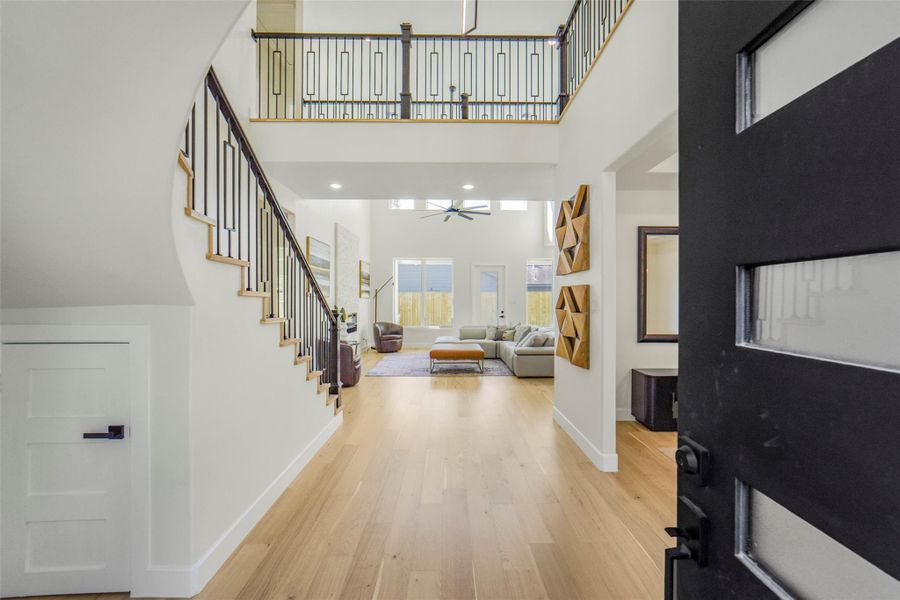 This inviting entryway features high ceilings, a modern staircase with elegant railing, and an open view into a spacious living area filled with natural light. The light wood flooring adds warmth, complemented by contemporary wall decor. Ideal for those seeking a blend of style and comfort.