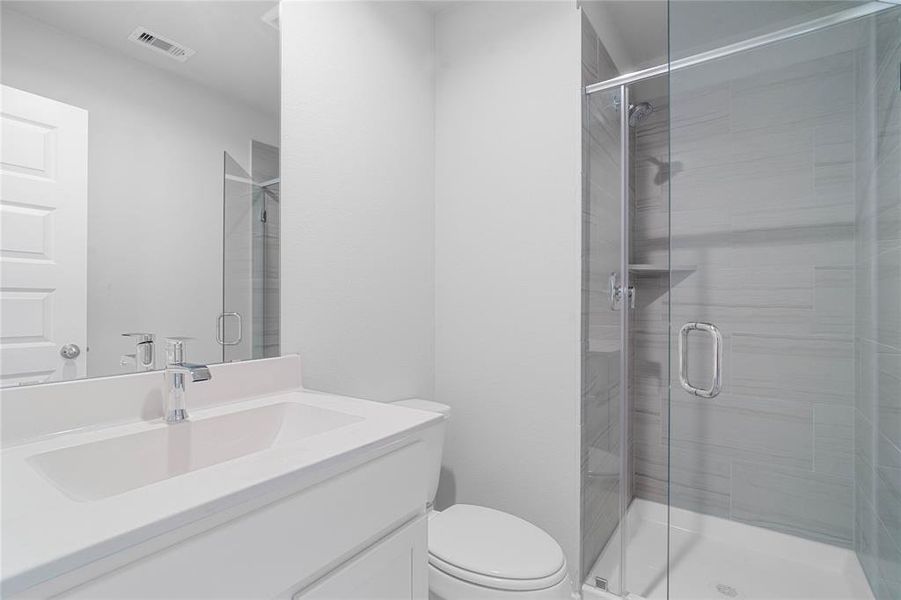 This private bath features tile flooring, shower with tile surround, light stained wood cabinets, beautiful light countertops, mirror, dark, sleek fixtures and modern finishes.