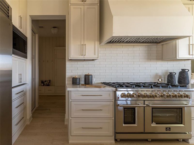 Kitchen featuring custom exhaust hood, appliances with stainless steel finishes, tasteful backsplash, light wood finished floors, and light stone counters
