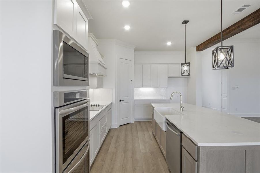 Dual tone kitchen featuring stainless steel appliances, light wood-style flooring, a kitchen island with sink, light stone countertops, and decorative light fixtures