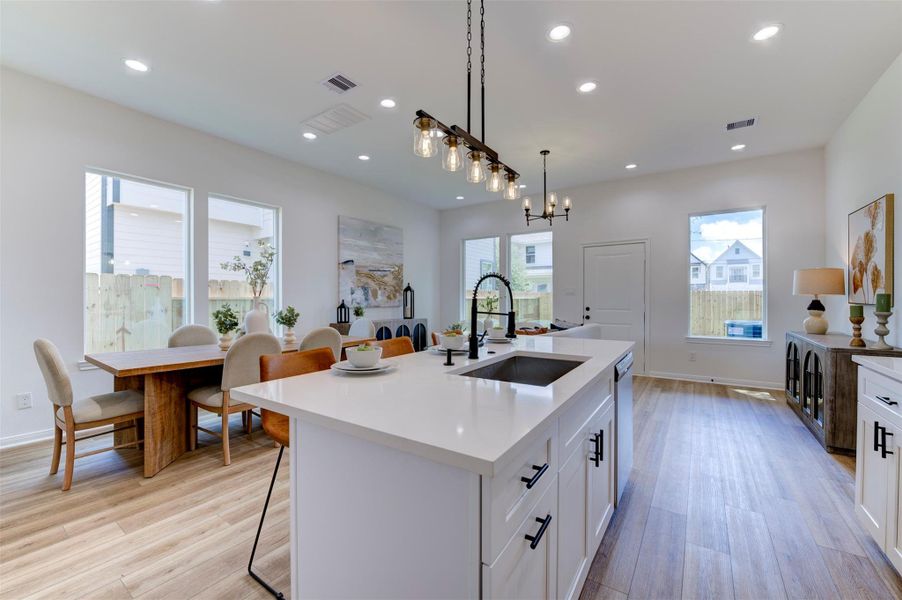 Wide view from kitchen island showing open flow to dining table, multiple large windows on three sides, matte black pendant lighting, hardwood floors, and rear exit door.