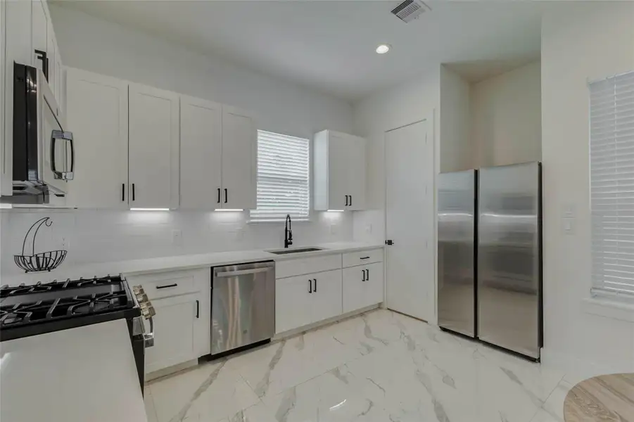 Kitchen detail view showcasing stainless appliances, white cabinetry, under-cabinet lighting, black fixtures, a window over the sink, and a full-size refrigerator. The crisp white palette, modern hardware, and marble-look floors create a polished, move-in-ready feel.