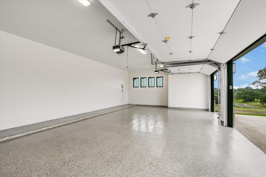 Spacious garage featuring a finished floor, white walls, multiple windows with black frames, and an open overhead door revealing exterior landscaping