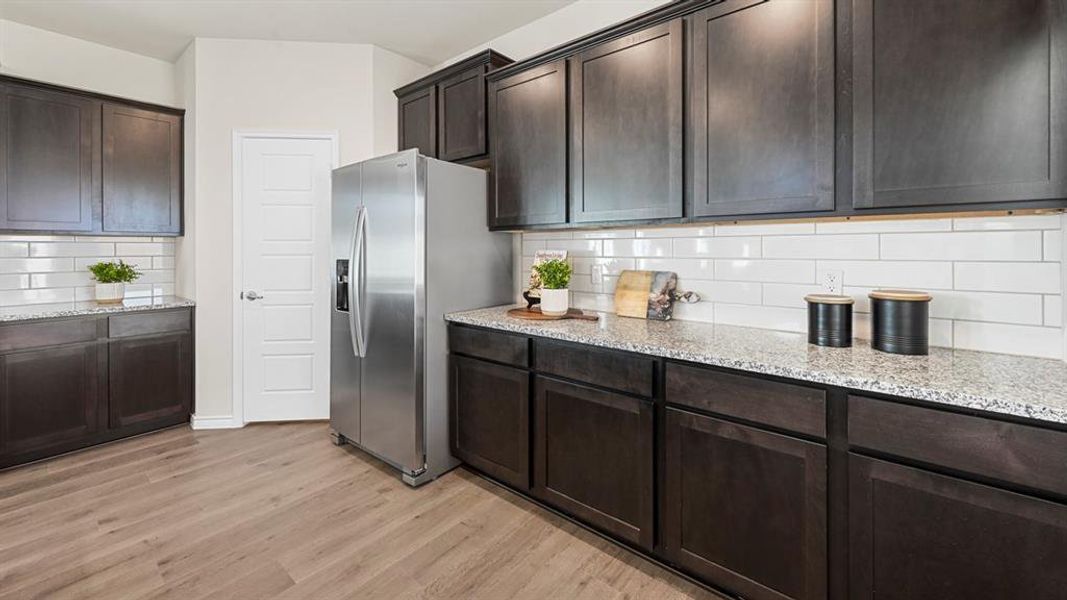 Kitchen featuring dark brown cabinets, stainless steel fridge with ice dispenser, light stone countertops, backsplash, and light wood-style floors Kitchen featuring dark brown cabinets, stainless steel fridge with ice dispenser, light stone countertops, backsplash, and light wood-style floors