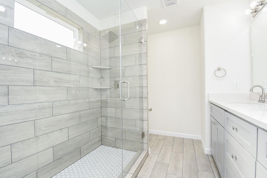 This is a modern bathroom featuring a large walk-in shower with gray wood-look tile, a frameless glass door, and built-in shelves. There's a white vanity with ample storage and a large mirror above it. The floor is finished with matching gray wood-look tiles. This is a modern bathroom featuring a large walk-in shower with gray wood-look tile, a frameless glass door, and built-in shelves. There's a white vanity with ample storage and a large mirror above it. The floor is finished with matching gray wood-look tiles.