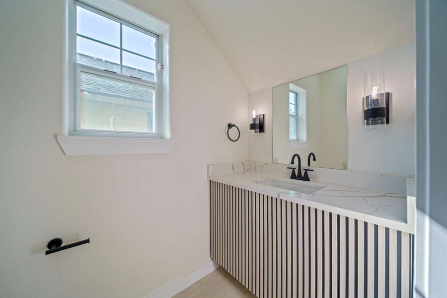 This bathroom features a modern design with a sleek, striped vanity and quartz countertop. It includes a large mirror, contemporary black fixtures, and is well-lit by wall sconces and natural light from a window. This bathroom features a modern design with a sleek, striped vanity and quartz countertop. It includes a large mirror, contemporary black fixtures, and is well-lit by wall sconces and natural light from a window.