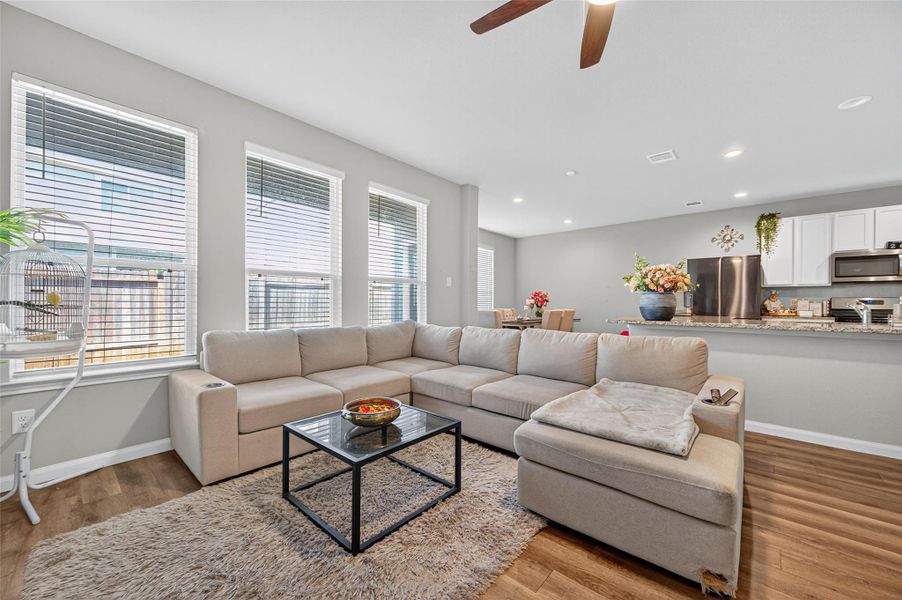 This photo showcases a modern, open-concept living area with a spacious beige sectional sofa, a sleek black coffee table, and a cozy rug. Large windows provide abundant natural light. The area seamlessly transitions into a stylish kitchen with a granite countertop, stainless steel appliances, and white cabinetry, offering a perfect blend of comfort and functionality.