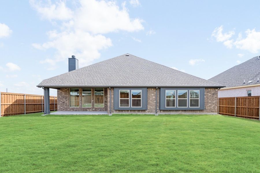 Rear view of brick home with covered patio, large windows, and fenced backyard with green lawn