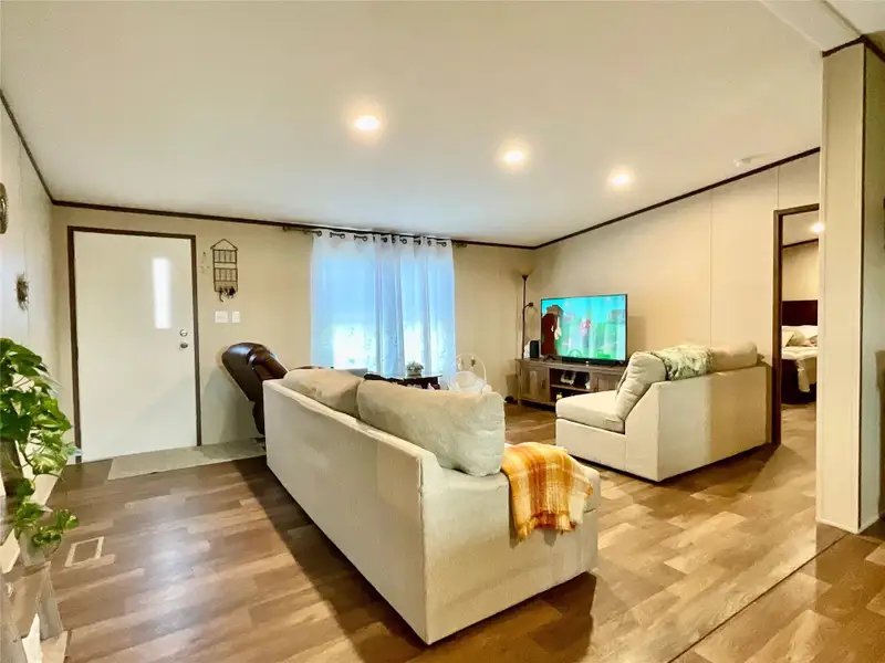 Spacious living area featuring wood-finish flooring, recessed lighting, a white entry door with a vertical window insert, and a large window with sheer white curtains