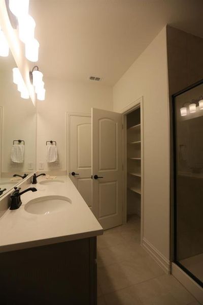 Bathroom featuring a dual vanity with white countertops and matte black fixtures, an open linen closet with shelving, and light-toned tile flooring