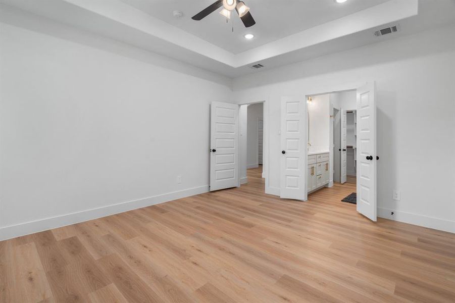 Unfurnished bedroom featuring a raised ceiling, ensuite bath, light wood-style flooring, a ceiling fan, and recessed lighting