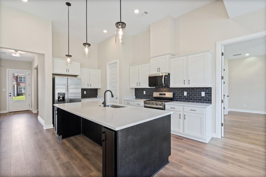 Kitchen featuring a central island with a light-toned countertop and dark cabinetry, stainless steel appliances, white upper and lower cabinetry, dark subway tile backsplash, and wood-finish flooring Kitchen featuring a central island with a light-toned countertop and dark cabinetry, stainless steel appliances, white upper and lower cabinetry, dark subway tile backsplash, and wood-finish flooring