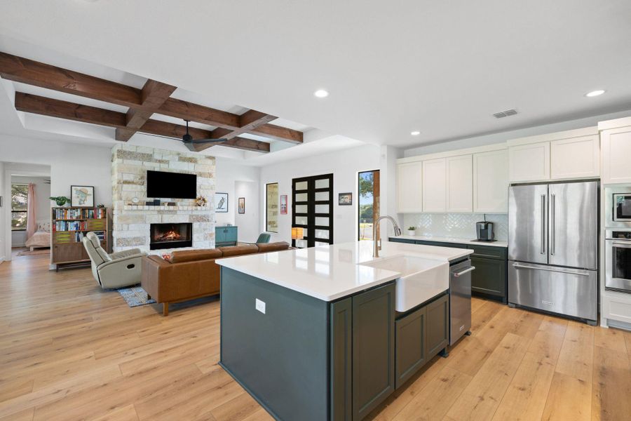 Kitchen featuring appliances with stainless steel finishes, coffered ceiling, white cabinets, a stone fireplace, and a center island with sink