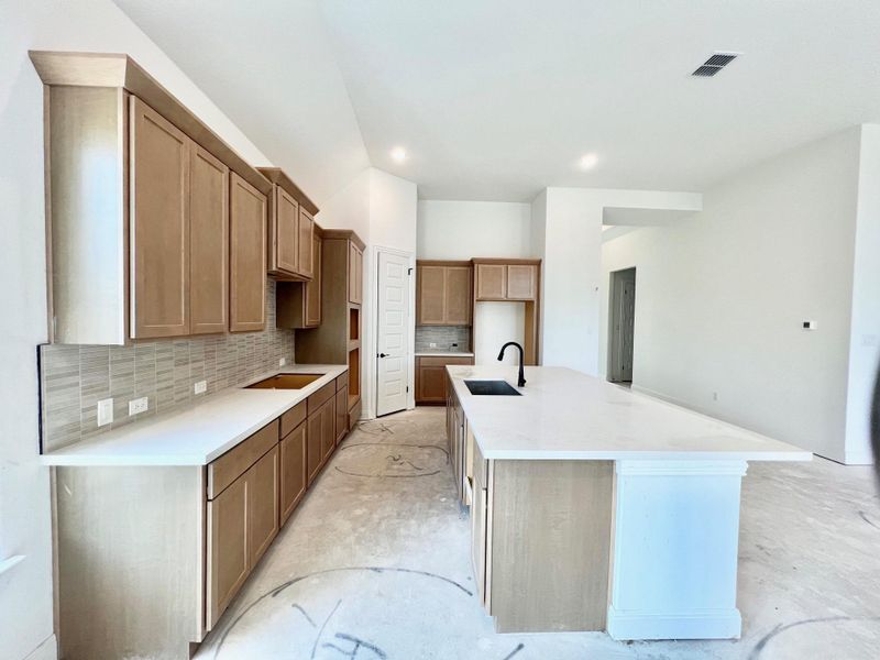 Kitchen with backsplash, light stone counters, a center island with sink, unfinished concrete flooring, and brown cabinetry Kitchen with backsplash, light stone counters, a center island with sink, unfinished concrete flooring, and brown cabinetry