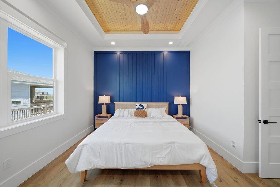 This bedroom features a contemporary design with a striking blue accent wall and wooden ceiling inset. It includes a comfortable bed with bedside tables and lamps, set on warm hardwood floors. A large window offers natural light and a view outside.