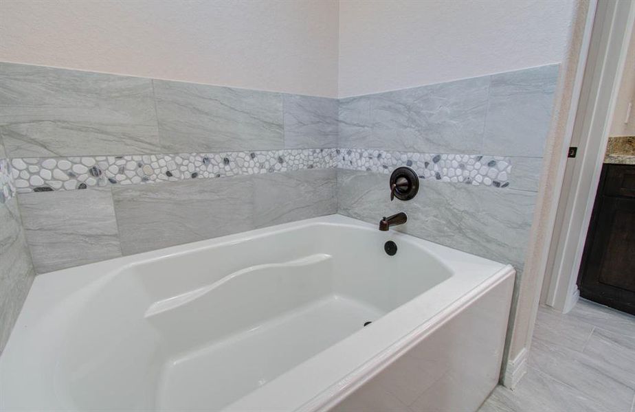 This is a modern bathroom featuring a built-in bathtub with gray tile surround and a decorative pebble tile accent. The hardware has a dark finish, and there is a glimpse of a wood cabinet, suggesting additional storage. The floor is finished with light-colored tiles.