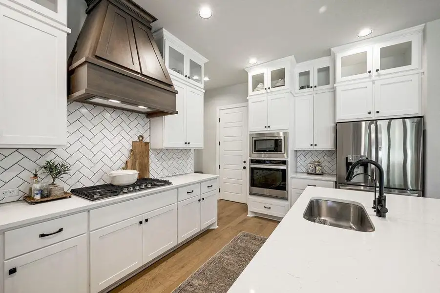 Kitchen featuring stainless steel appliances, custom range hood, glass insert cabinets, wood finished floors, and white cabinets Kitchen featuring stainless steel appliances, custom range hood, glass insert cabinets, wood finished floors, and white cabinets