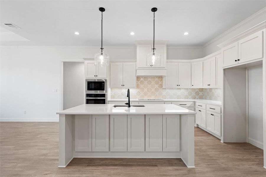 Kitchen with ornamental molding, white cabinets, tasteful backsplash, a kitchen island with sink, and light wood-style flooring Kitchen with ornamental molding, white cabinets, tasteful backsplash, a kitchen island with sink, and light wood-style flooring