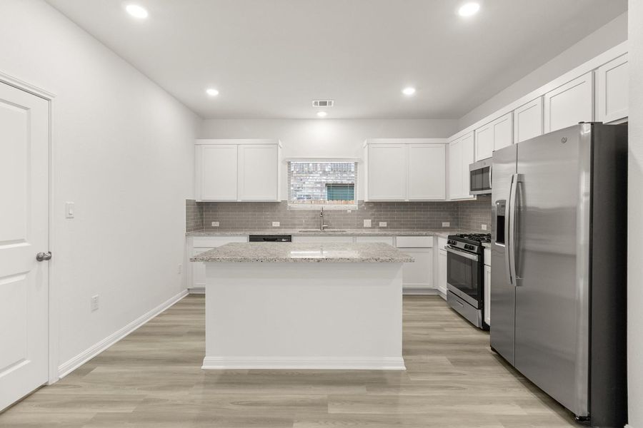 The kitchen features an island with a light-colored countertop, white cabinetry, and stainless steel appliances, including a refrigerator, microwave, and range