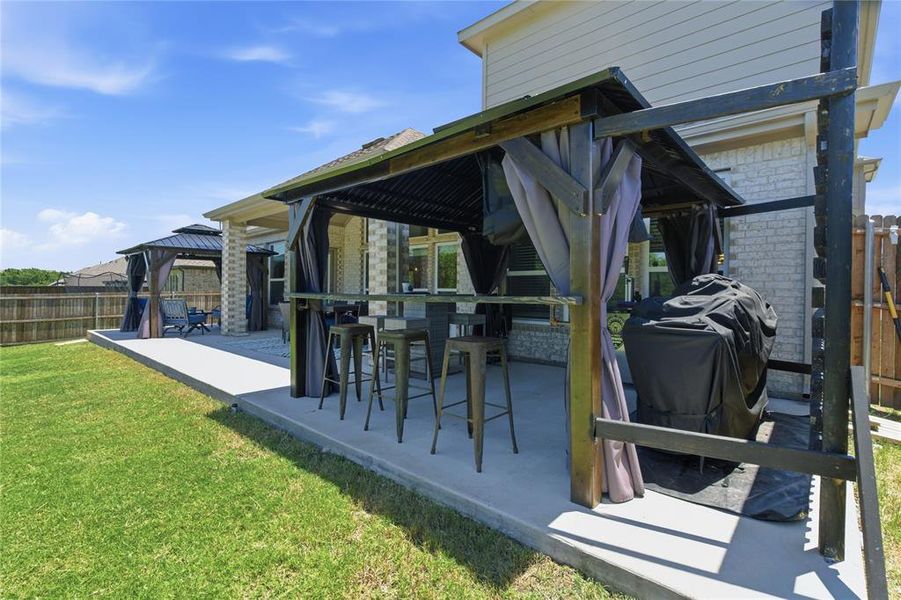 Expansive concrete patio featuring two covered pergola structures, a built-in outdoor bar area with seating, and a perimeter wood privacy fence