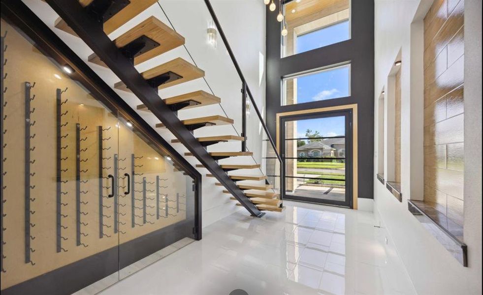 This is a modern entryway featuring a floating wooden staircase with a sleek black metal railing. The wall is equipped with a wine storage display, and the area is well-lit with natural light streaming in through large windows, creating an inviting and contemporary space.