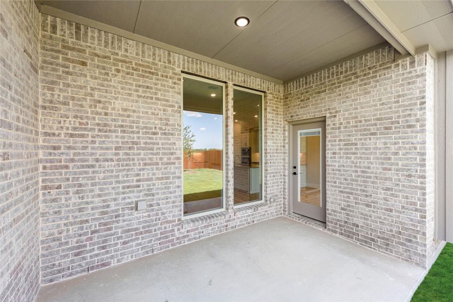 Covered back patio with concrete floor, brick exterior wall, rear entry door, and large window overlooking fenced backyard.