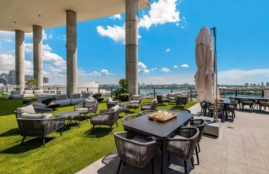 Expansive covered terrace featuring structural concrete columns, synthetic turf surface, stone-tile flooring, and a glass panel perimeter railing Expansive covered terrace featuring structural concrete columns, synthetic turf surface, stone-tile flooring, and a glass panel perimeter railing