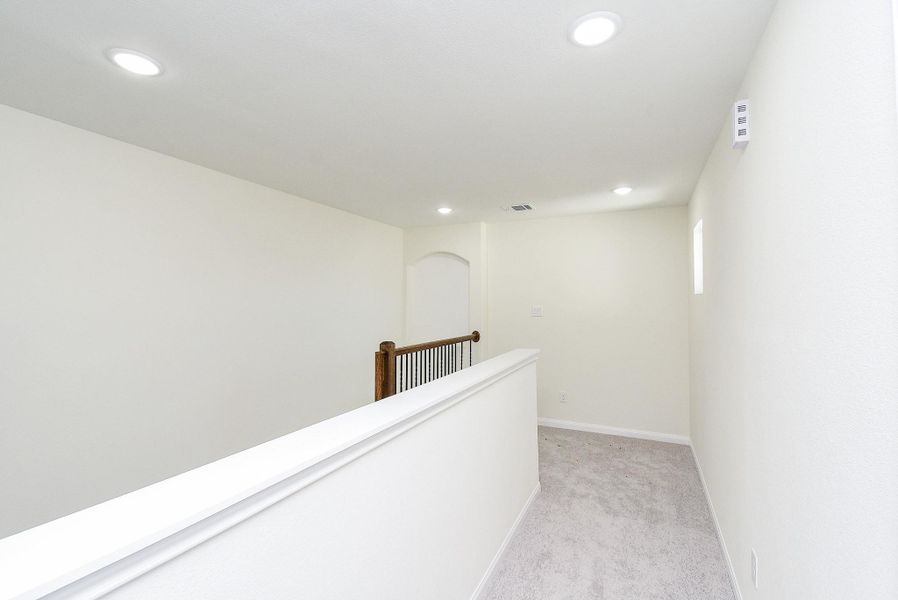 Bright, empty hallway with carpeted floor, white walls, recessed lighting, and a wooden banister on the upper level of a modern home.