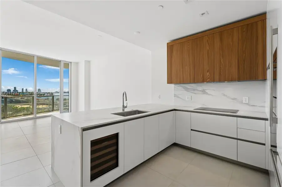 Kitchen featuring an L-shaped layout, wood-finish upper cabinetry, white flat-panel lower cabinetry, integrated stone-finish countertops and backsplash, and an under-counter wine refrigerator