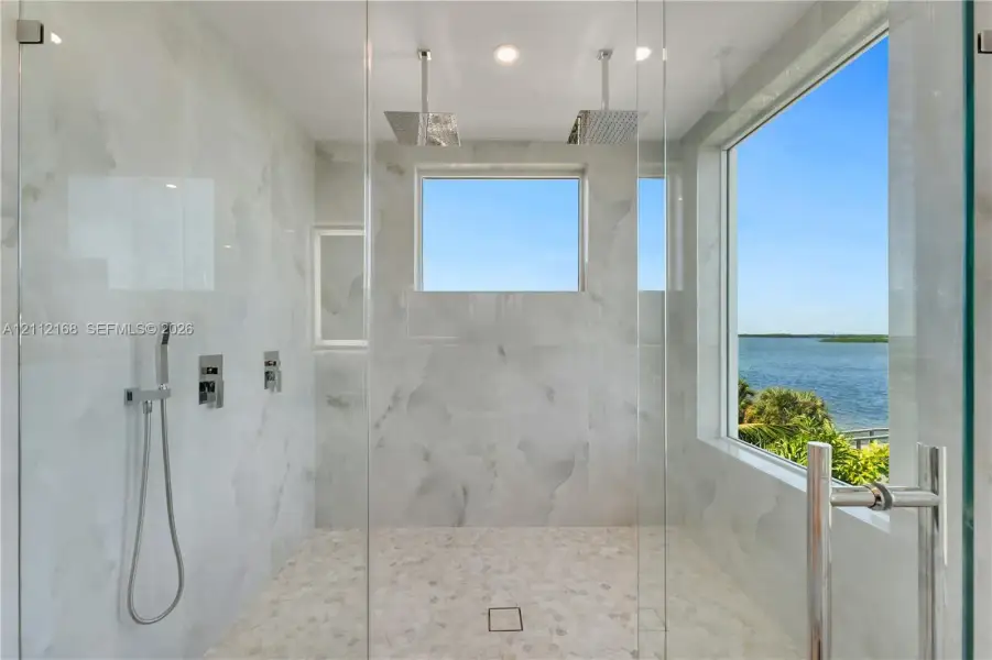 Walk-in shower featuring floor-to-ceiling stone-patterned tile, dual ceiling-mounted rainfall showerheads, a handheld sprayer, a glass enclosure, and a large window Walk-in shower featuring floor-to-ceiling stone-patterned tile, dual ceiling-mounted rainfall showerheads, a handheld sprayer, a glass enclosure, and a large window
