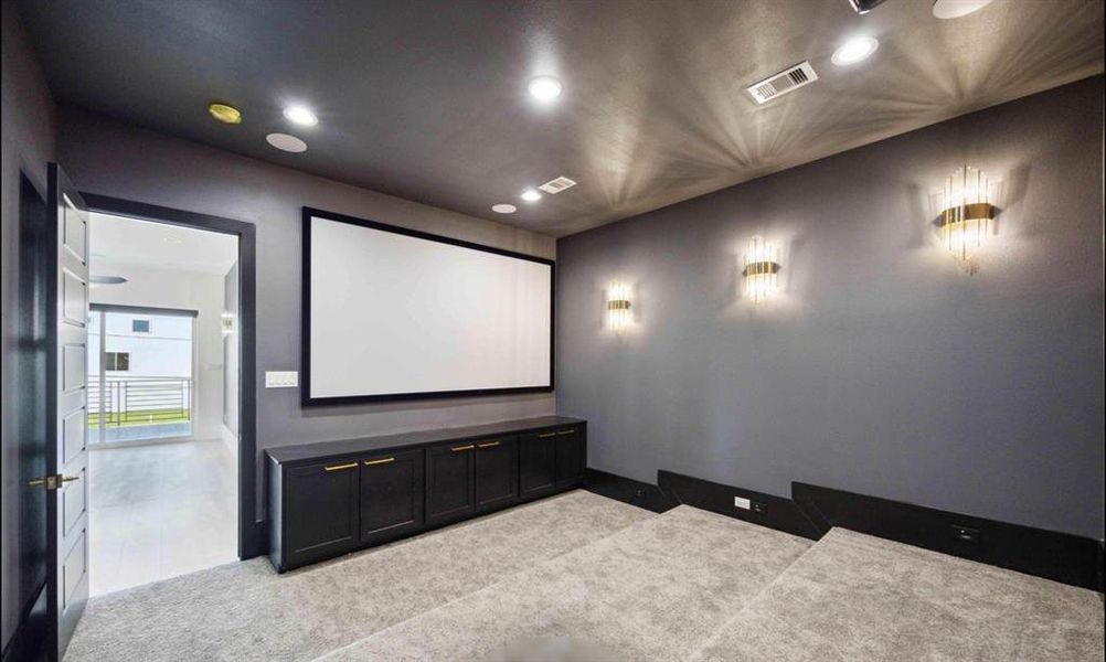 This is a modern home theater room featuring dark grey walls, recessed lighting, and elegant wall sconces. A large projection screen is mounted on the main wall, with sleek black cabinetry below. The room is carpeted and opens to a bright space through double doors, suggesting a private yet accessible entertainment area within the home.