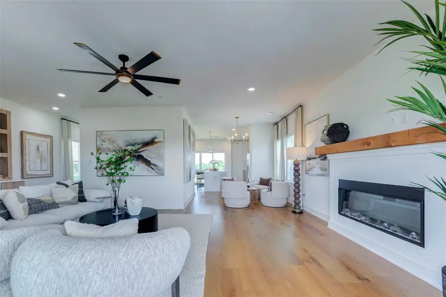 This expansive living area offers a clear view through the home, highlighting the seamless connection between the lounge spaces, dining area, and kitchen. A linear fireplace with a warm wood mantel anchors one side of the room, while custom built-ins define the other. Wide-plank floors, contemporary ceiling fans, recessed lighting, and soft neutral furnishings create a cohesive setting designed for comfortable everyday living and easy entertaining.