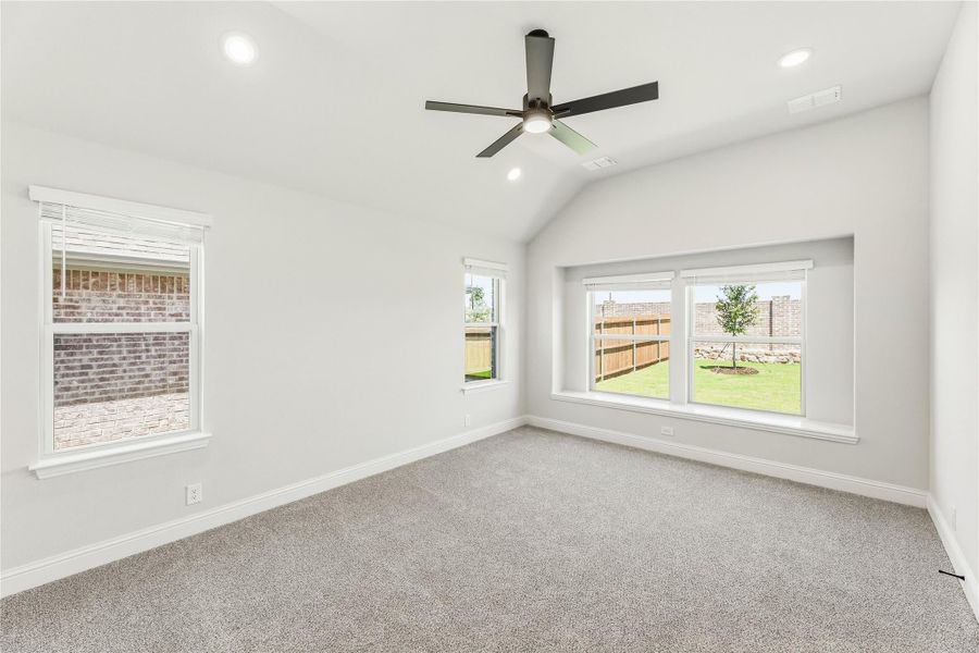 Empty bedroom with vaulted ceiling, ceiling fan, carpet flooring, and multiple windows overlooking backyard