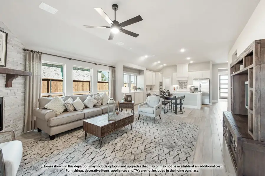 Open-concept living room with stone fireplace, ceiling fan, and hardwood floors leading to kitchen