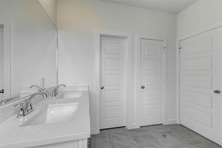 Primary bath vanity area featuring dual undermount sinks, quartz-style countertop, chrome faucets, large mirror, tile flooring, and nearby doorways for closet and bath access. The bright white cabinetry and neutral finishes create a clean, functional space for everyday routines.