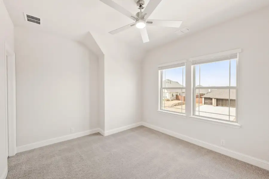 Empty bedroom with vaulted ceiling, ceiling fan, gray carpet, and two windows overlooking neighborhood